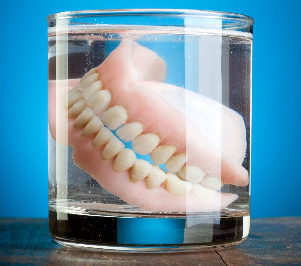 a set of traditional dentures in a glass of water. there is a dark blue background behind the glass. 